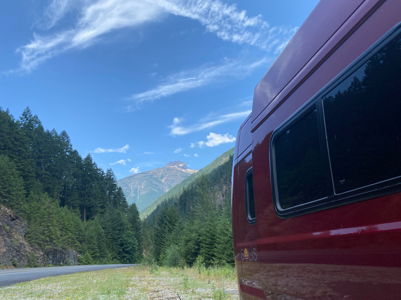 Scenic mountain road trip: A red RV rental parked by lush forests and a distant peak under a blue sky.
