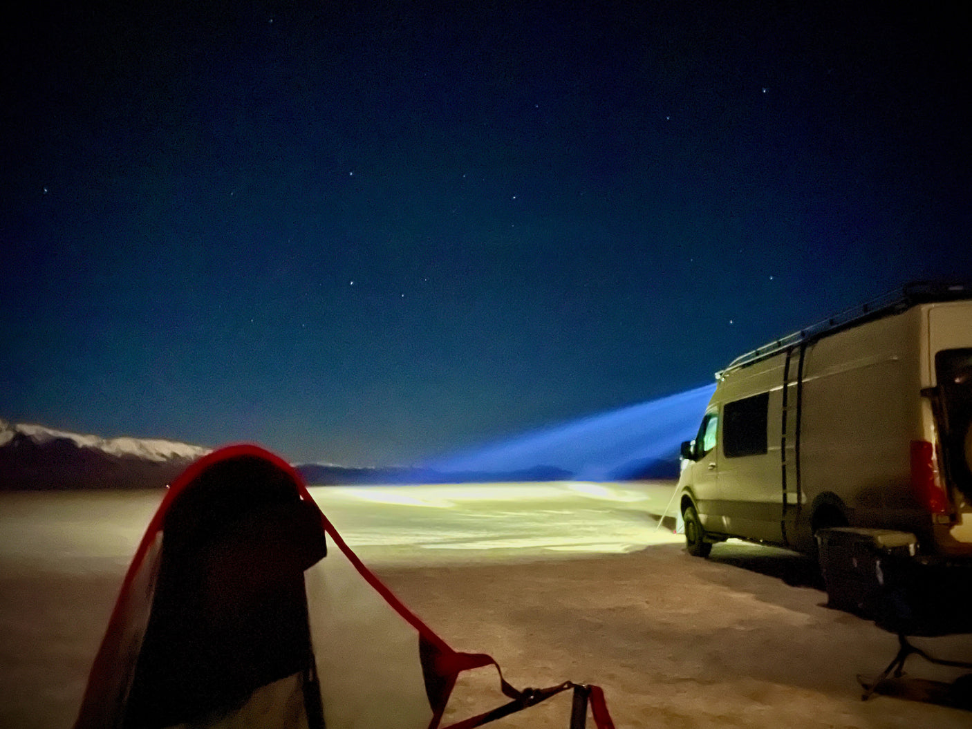 Night sky camping: an RV rental illuminates the vast landscape beside a red tent, with distant snowy mountains under the stars.