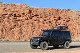 Black Jeep Wrangler with roof tent, an adventure RV rental, parked roadside against towering red rock formations under blue sky.