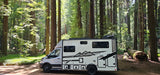 White camper van RV rental parked in a vibrant redwood forest. Sunbeams pierce the canopy, illuminating tall trees and the forest floor.