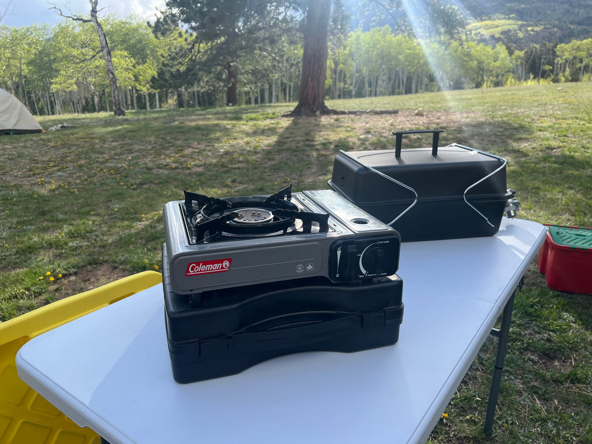 Campsite cooking gear: Coleman stove & grill on a table, with a tent and trees. Ideal for an RV rental adventure! — photo 11
