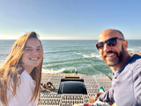 Smiling couple on their RV rental rooftop, enjoying vast ocean views and the coastal breeze, with a person by the waves below.