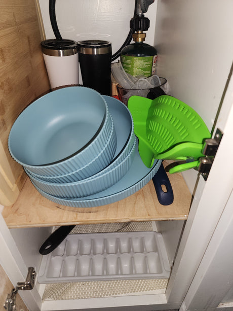 Inside an RV rental cabinet: stacked blue dishes, a green propane tank, travel mugs, and a bright green silicone strainer.