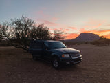 Dark SUV on a dirt road at sunset by mountains & trees, driver door open. An outdoor adventure, perfect for an rv rental getaway.