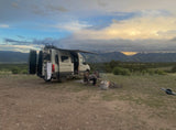 RV rental camper van parked in mountains at dusk. Person by campfire; doors open, Onewheel nearby. Amazing outdoor escape!