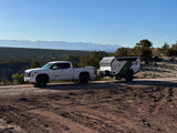 White pickup towing a TETON off-road RV rental on a dirt trail. Scenic desert mountains for your next adventure camping trip.