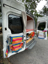 Open white camper van rear, revealing a bed, stored gear, and door-mounted beach chairs/drying rack. Great for an RV rental trip.