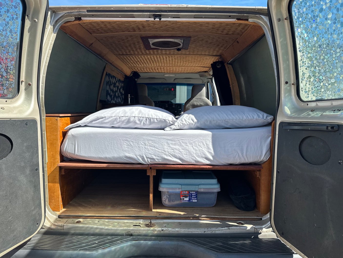 Custom camper van interior, perfect for an rv rental, showing a comfy bed, under-bed storage, and unique patterned windows.