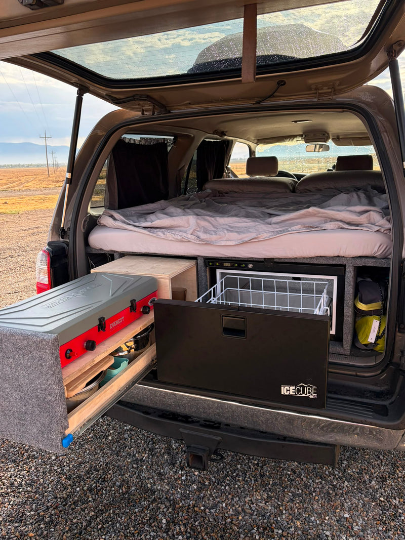Camper setup deployed at a desert campsite — bed made on the platform, pull-out cooking surface with two-burner stove and fridge drawer open — photo 3