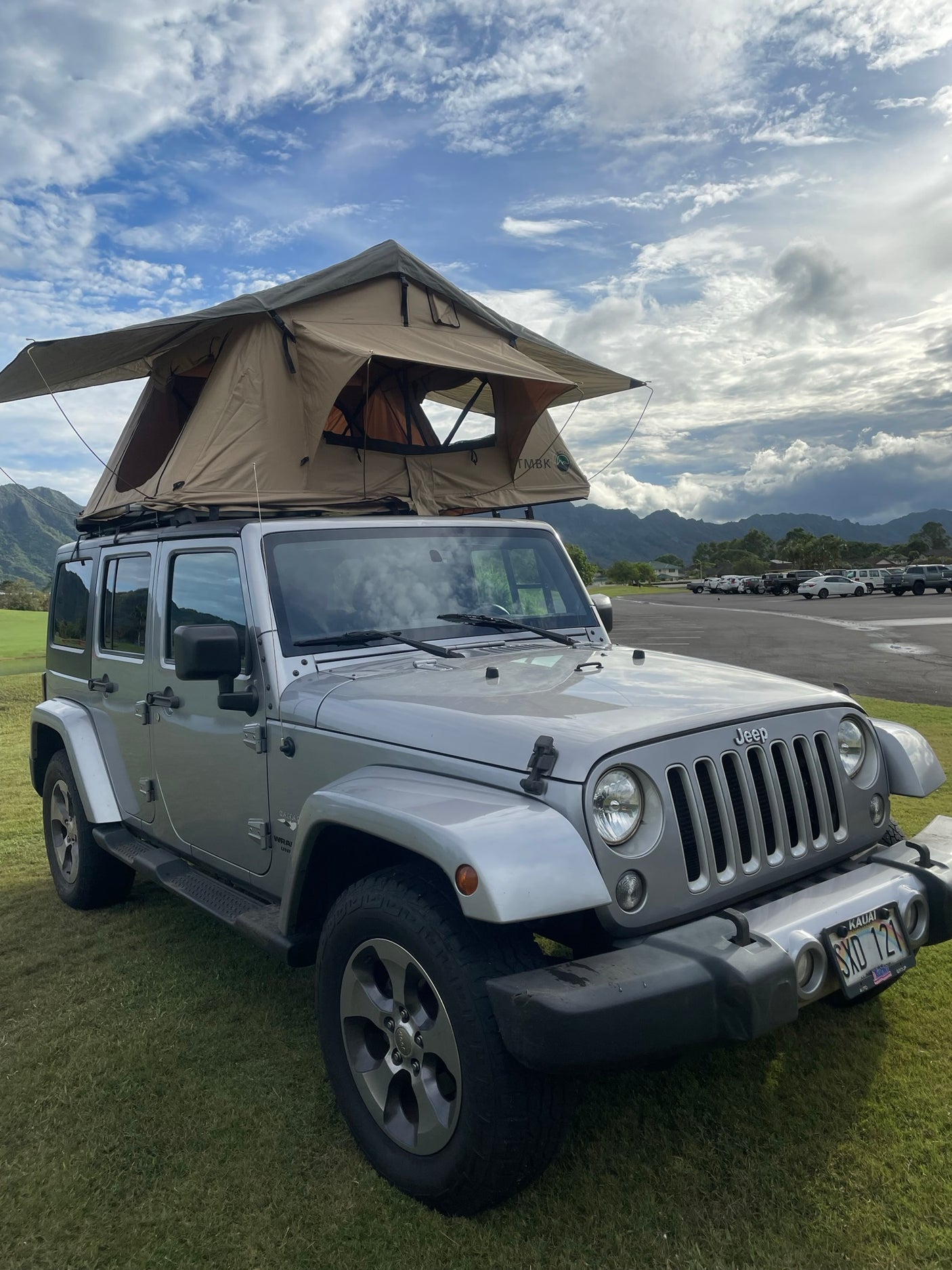 Silver Jeep with a deployed rooftop tent on a grassy field, mountains in background. An RV rental for rugged Kauai outdoor adventures.