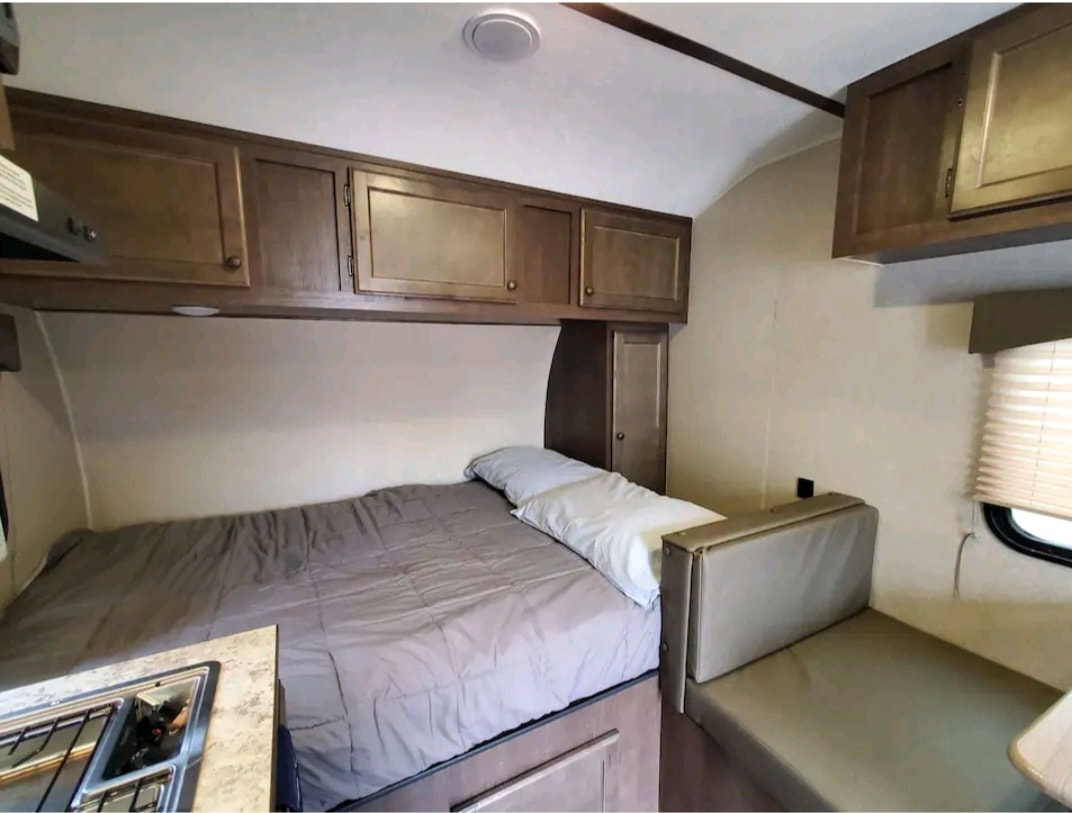 Cozy RV rental bedroom setup with a bed, overhead wood cabinets, a compact kitchenette, and a sofa/dinette area. — photo 11