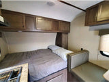 Cozy RV rental bedroom setup with a bed, overhead wood cabinets, a compact kitchenette, and a sofa/dinette area.