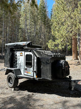 An Off Grid Expedition 2.0 camper trailer, like an RV rental, parked in a sunny forest setting.