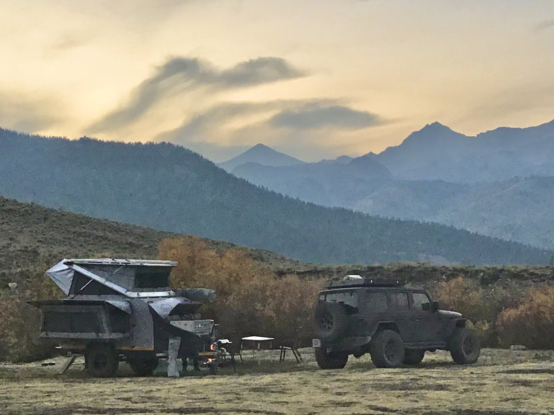 Dusk camp in an open mountain valley with the rooftop tent raised, awning out and a campfire burning beside the trailer, layered ridgelines behind — photo 5