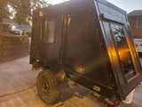 Side view of a compact, black teardrop camper, possibly an rv rental. It features a side door, a rear window, and one visible wheel.