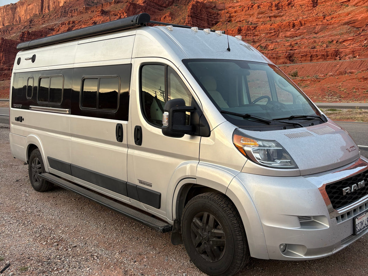 A silver RV rental camper van, possibly a Winnebago Travato, parked on gravel in front of towering red rock cliffs, ready for travel. — photo 1