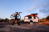 Person carrying a bike on rocky terrain next to a white RV rental camper, set up for outdoor adventure.