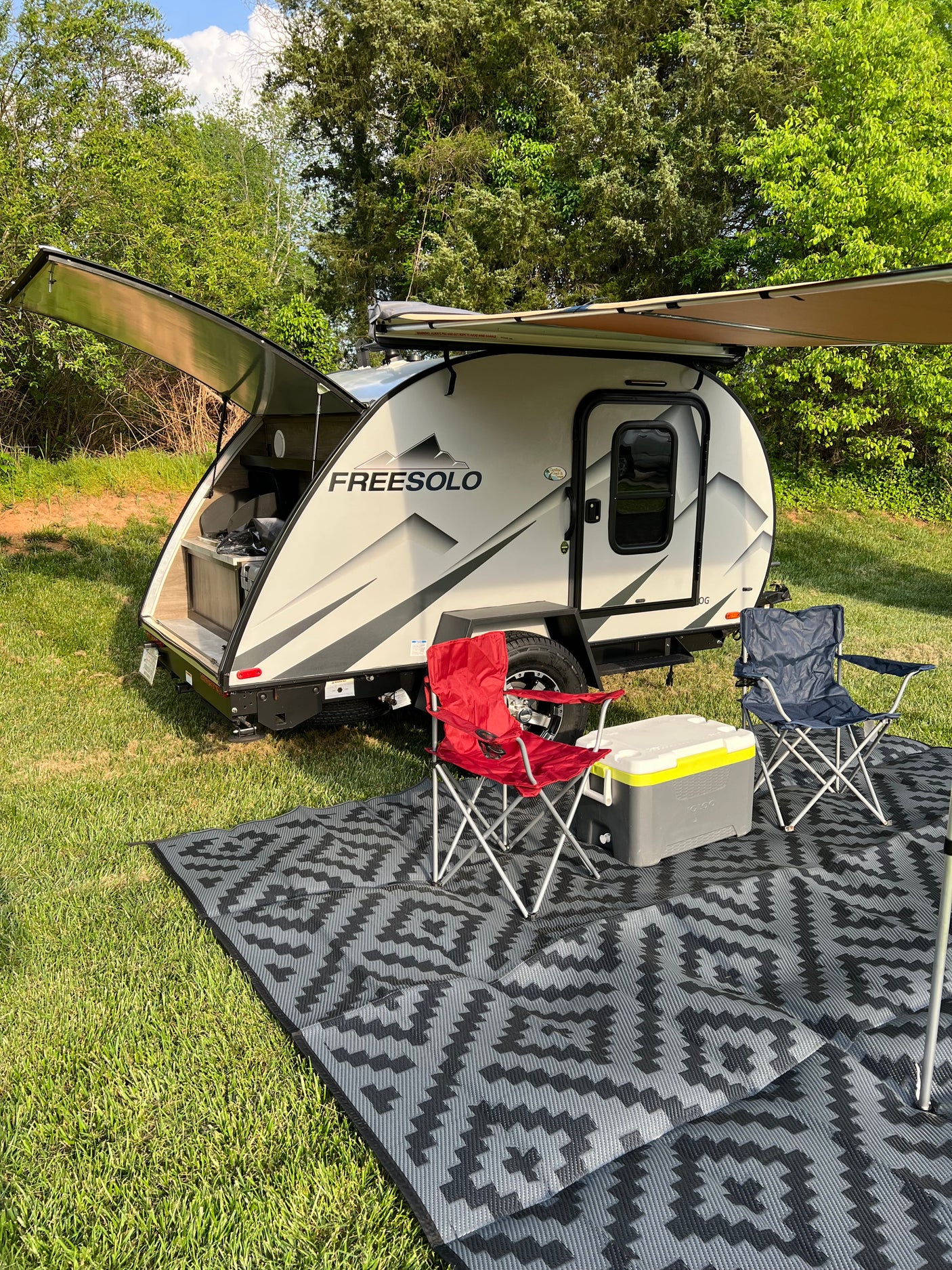RV rental: Modern teardrop travel trailer with extended awning, outdoor kitchen, chairs, & cooler on a patterned mat in a grassy area.