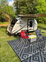 RV rental: Modern teardrop travel trailer with extended awning, outdoor kitchen, chairs, & cooler on a patterned mat in a grassy area.