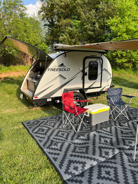 RV rental: Modern teardrop travel trailer with extended awning, outdoor kitchen, chairs, & cooler on a patterned mat in a grassy area.