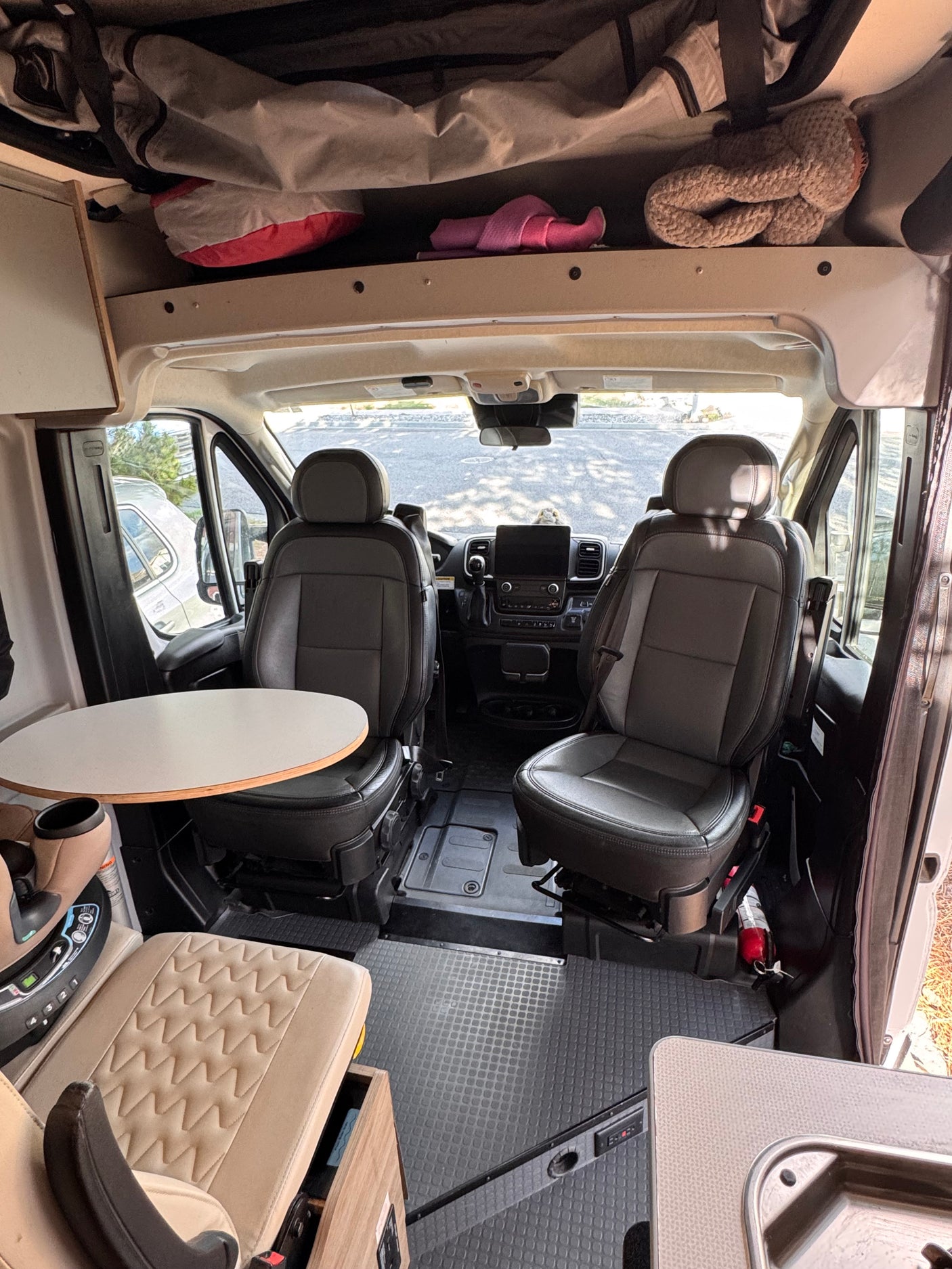 Interior of an RV rental camper van. Front cabin with swiveling driver and passenger seats, a small dining table, and overhead storage.