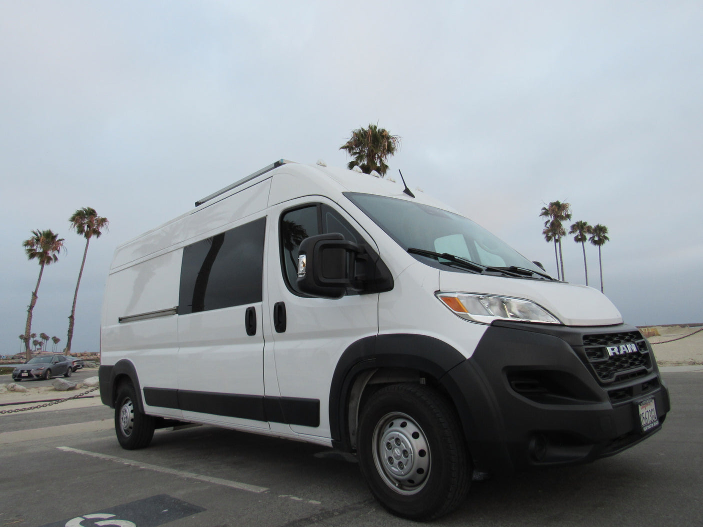 White Ram ProMaster RV rental van parked at a beach with palm trees under a cloudy sky.
