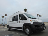 White Ram ProMaster RV rental van parked at a beach with palm trees under a cloudy sky.