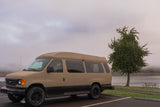 Tan Ford E-Series camper van (RV rental) parked in a lot beside a foggy lake with a tree & picnic area.