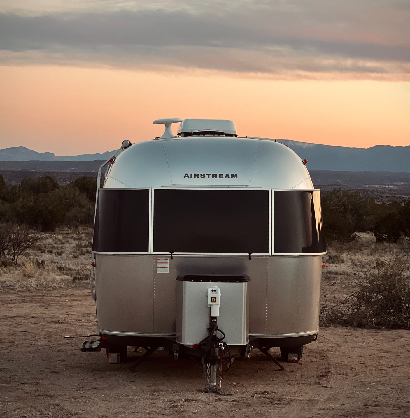 An Airstream RV rental from behind, parked in a rugged desert landscape at sunset with distant mountains.