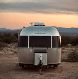 An Airstream RV rental from behind, parked in a rugged desert landscape at sunset with distant mountains.