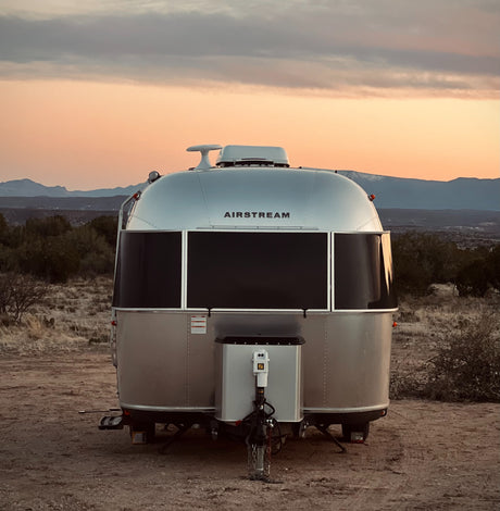 An Airstream RV rental from behind, parked in a rugged desert landscape at sunset with distant mountains.