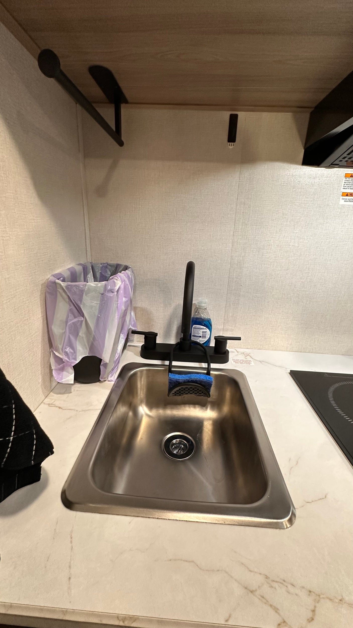This RV rental kitchen features a modern sink, black faucet, soap, sponge, trash can, and clean marble-look countertop with a cooktop.