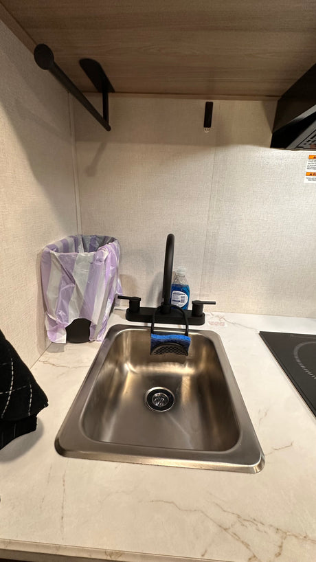 This RV rental kitchen features a modern sink, black faucet, soap, sponge, trash can, and clean marble-look countertop with a cooktop.