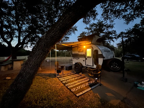 Cozy night camping with an RV rental. A lit Sunray travel trailer, awning, rug, & chairs under trees.
