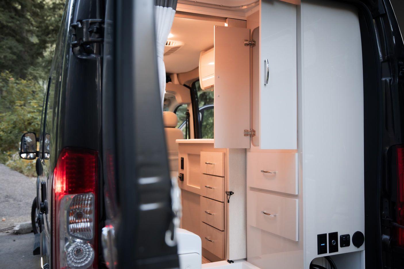 Inside an RV rental: a bright camper van interior with modular storage, light wood and white cabinetry, seen from the rear.