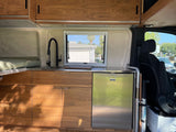 RV rental interior: A modern wooden kitchenette with a sink, black faucet, stainless refrigerator, and window.