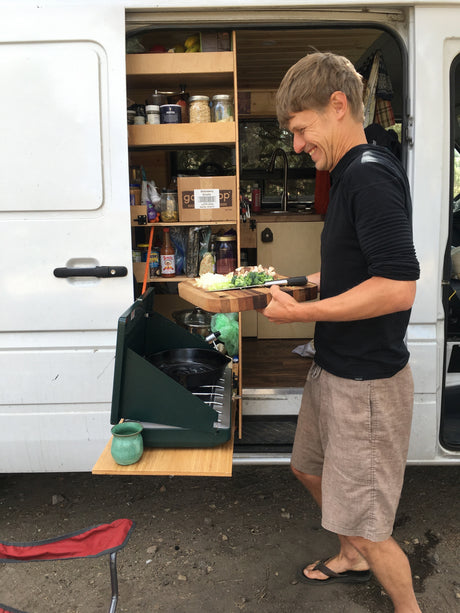 A man smiles while prepping veggies on a cutting board outside an RV rental, cooking on a portable stove next to his camper van kitchen.
