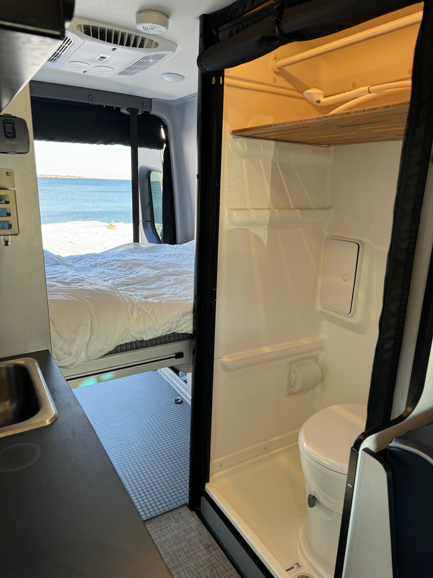 RV rental interior showing bed with beach view, wet bath with toilet/shower, and kitchenette.