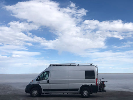 Silver camper van, an RV rental, with bikes on a rack, parked on vast salt flats under a cloudy blue sky.