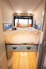 A comfortable, made bed with soft bedding and pillows inside an RV rental, featuring mugs and a book, with windows showing outdoor scenery.