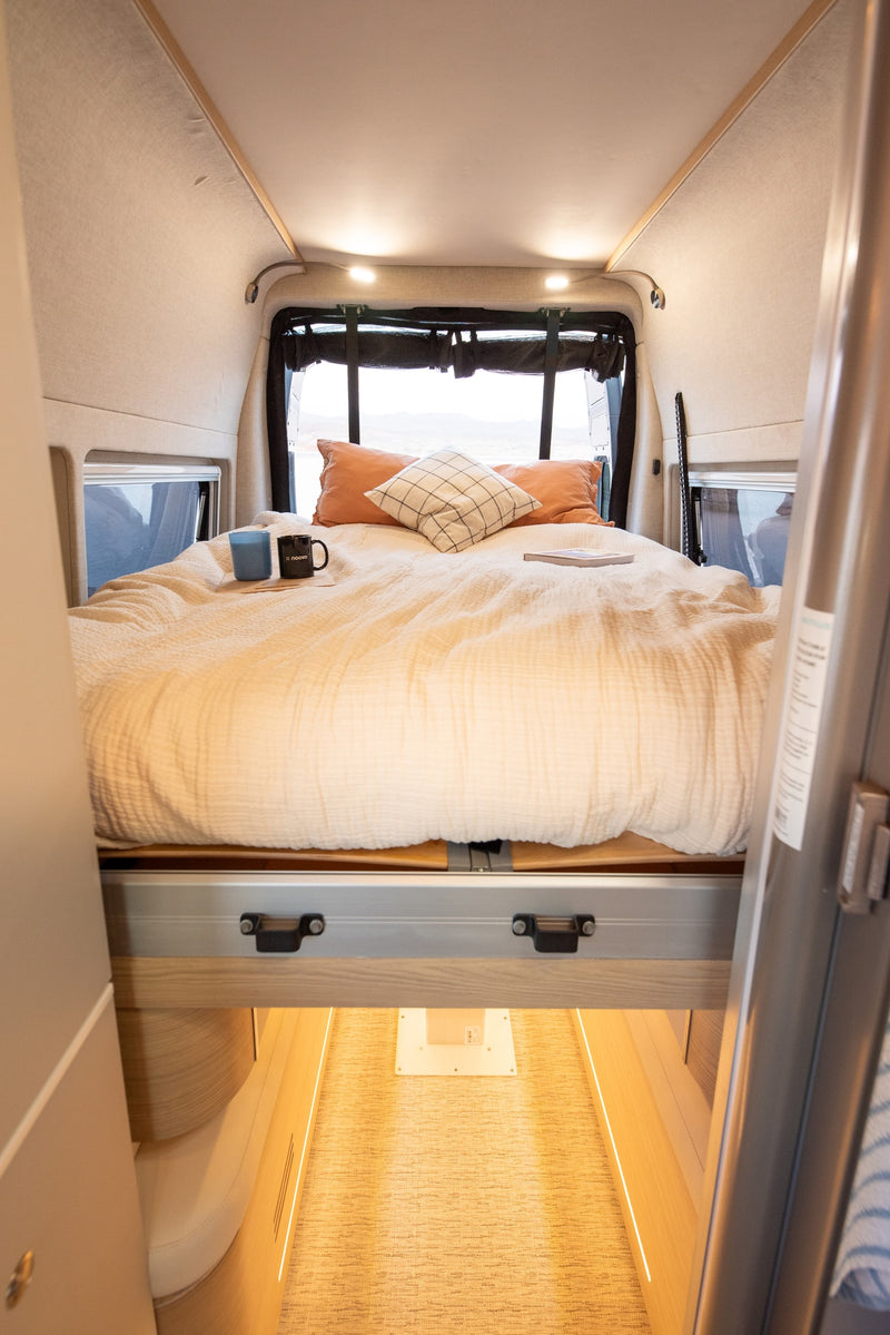 A comfortable, made bed with soft bedding and pillows inside an RV rental, featuring mugs and a book, with windows showing outdoor scenery. — photo 11