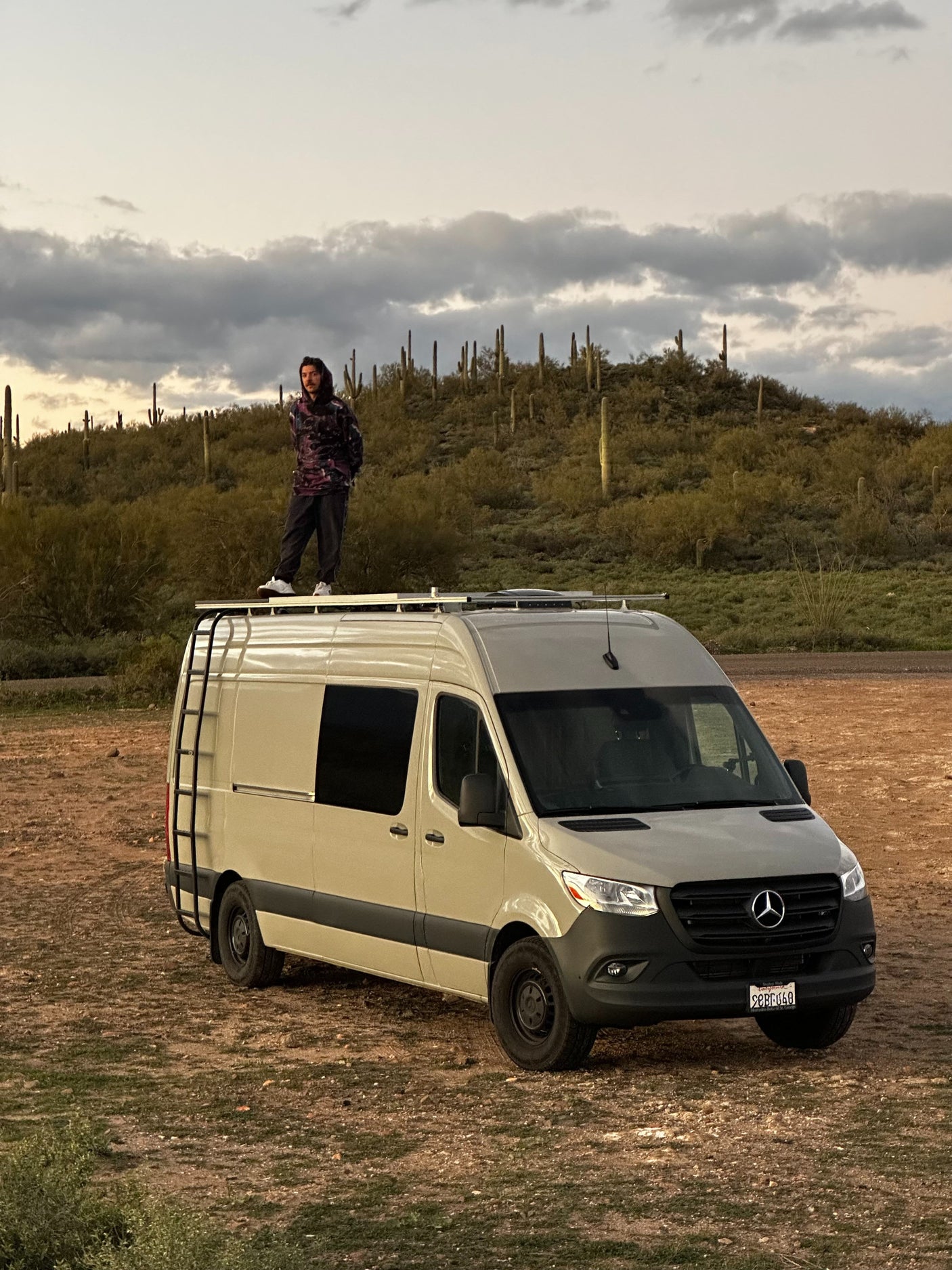 A person stands atop a Mercedes Sprinter rv rental amidst a desert landscape with saguaro cacti under a cloudy sky.
