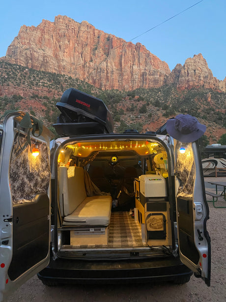 Cozy, customized camper van with open doors and lights, backed by red mountains. Great rv rental for your next adventure!