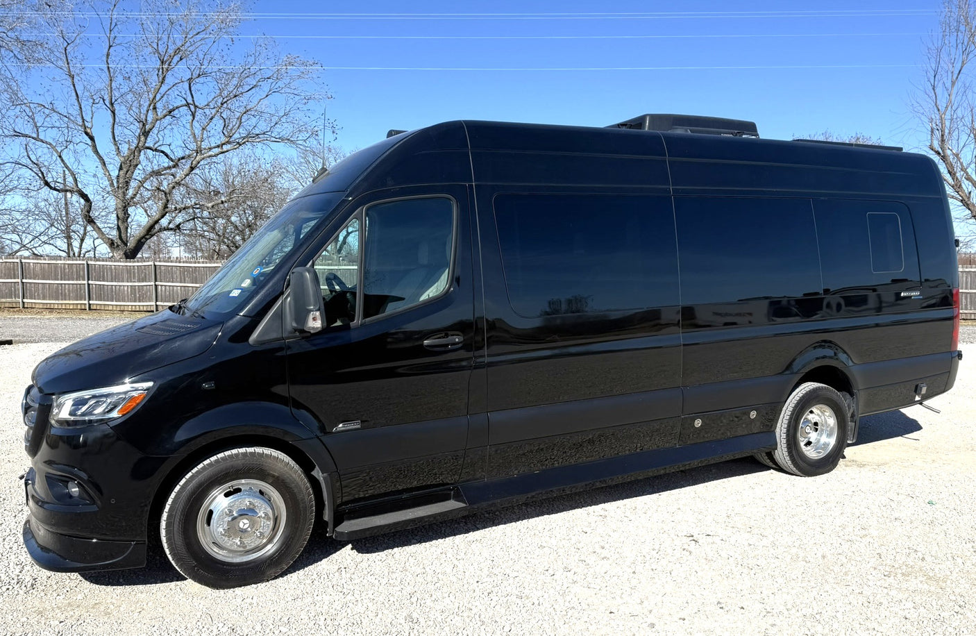 Black Sprinter-style RV rental van, parked on gravel in daylight with trees in background, ready for adventure.