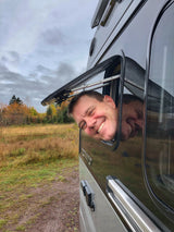 Smiling man and his reflection peek from an RV rental window in a vibrant fall landscape.