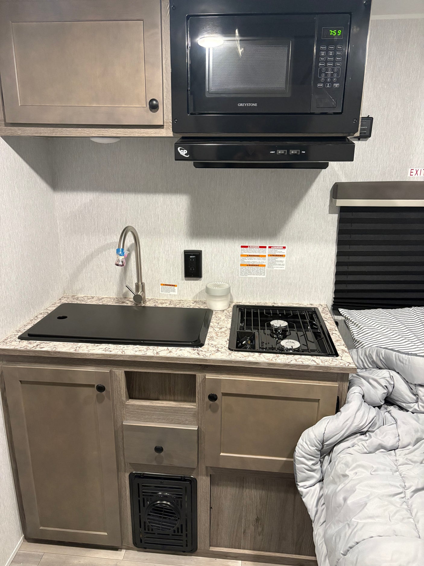 This RV rental galley features a Greystone microwave (7:59), sink, two-burner stove, cabinets, and adjacent bed, showcasing a functional compact space.
