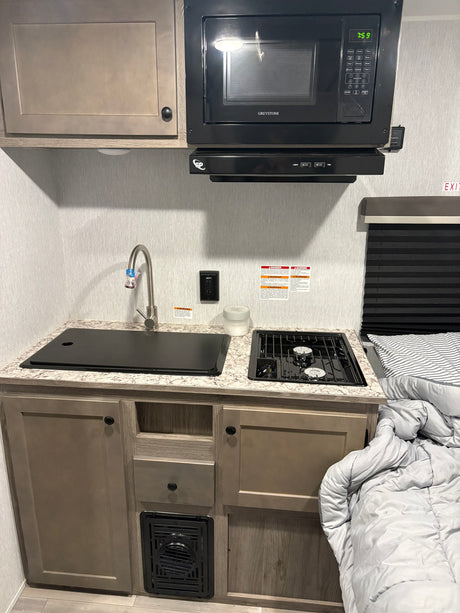 This RV rental galley features a Greystone microwave (7:59), sink, two-burner stove, cabinets, and adjacent bed, showcasing a functional compact space.