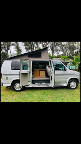 Silver pop-top camper van parked on grass with its side door open, revealing a compact interior for RV rental.