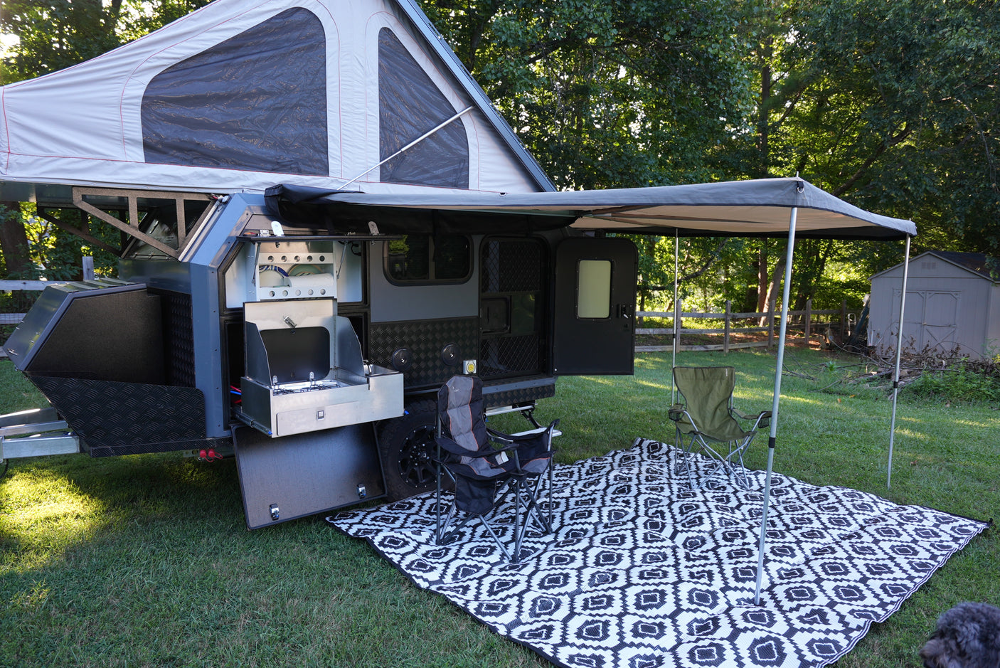 This RV rental setup shows a pop-top camper with an outdoor kitchen, awning, chairs, and mat on a grassy lawn.