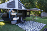 This RV rental setup shows a pop-top camper with an outdoor kitchen, awning, chairs, and mat on a grassy lawn.